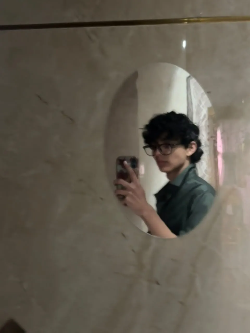 Aayan in glasses and a green shirt, taking a mirror selfie in an oval mirror