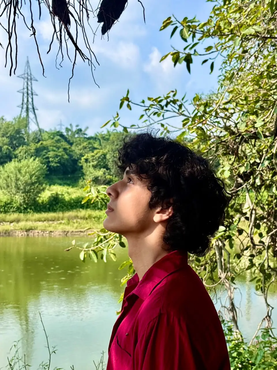 Aayan in a red shirt by a lake, looking up at the trees