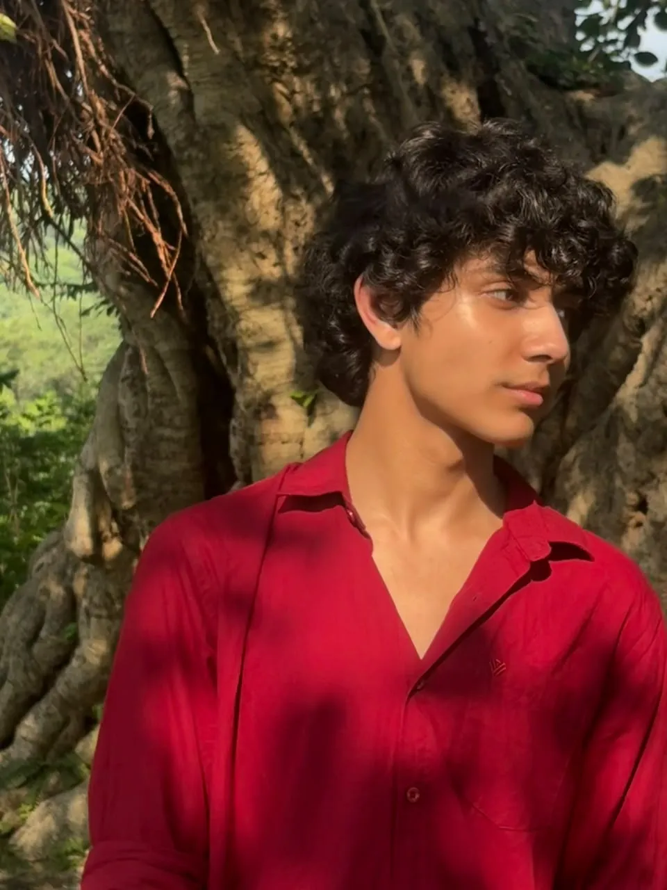 Aayan in a red shirt against a large tree trunk, dappled sunlight