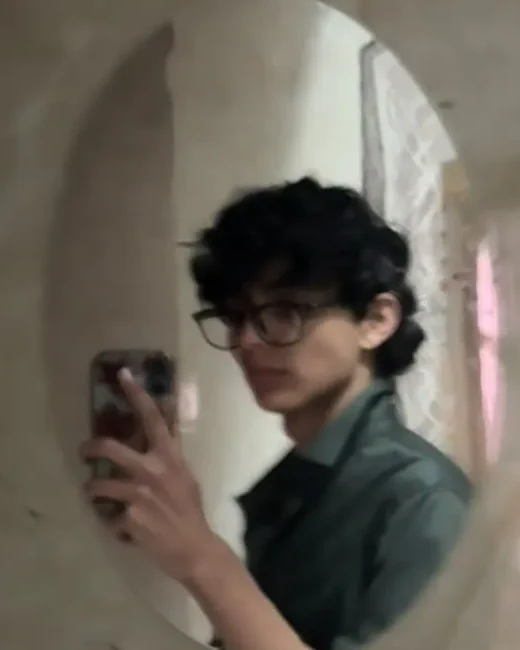 Aayan in glasses and a green shirt, taking a mirror selfie in an oval mirror